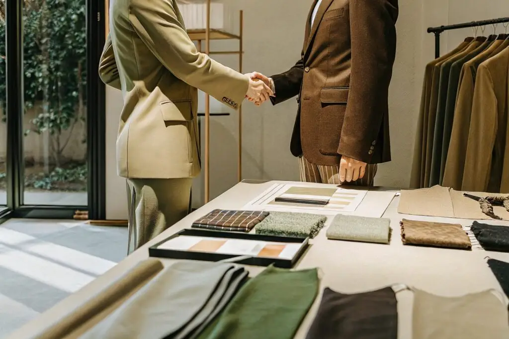 A clothing manufacturer meeting with an American fashion brand representative, sealing a deal with a handshake over a table filled with fabric samples and design materials.