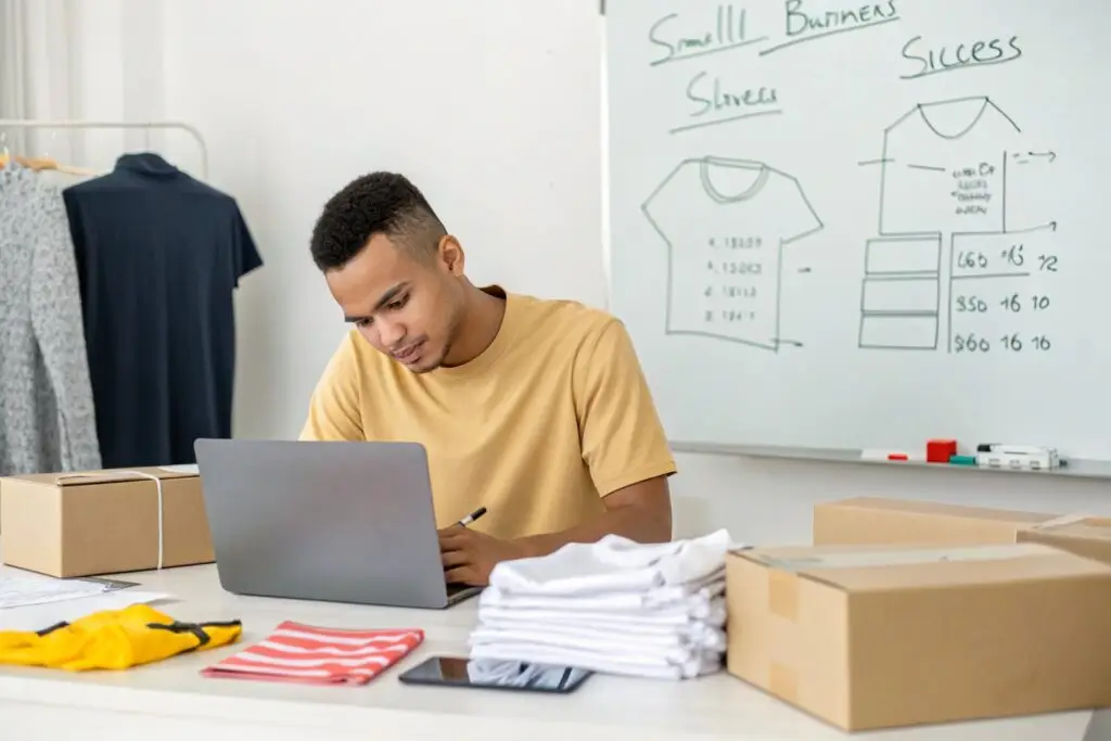 Young clothing business owner working on laptop beside product boxes