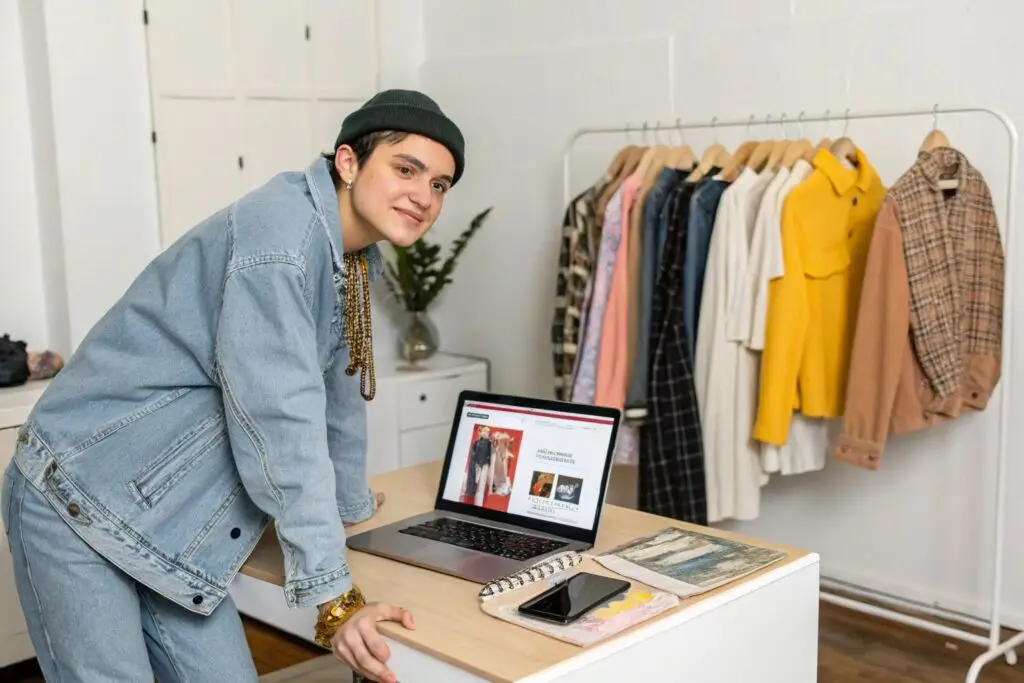 Smiling person in denim outfit standing next to a laptop displaying fashion items