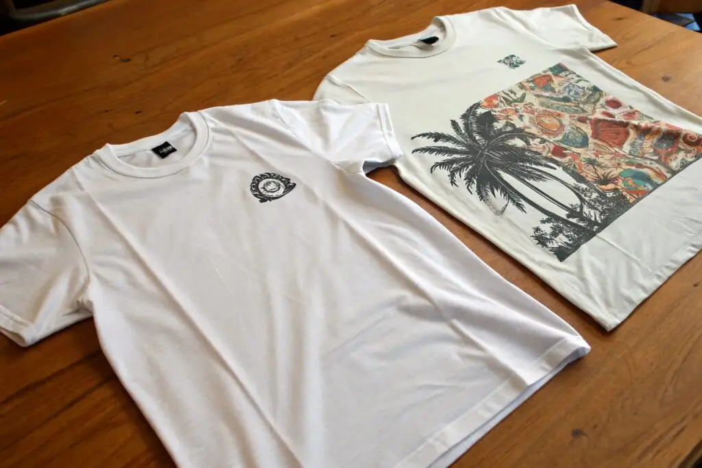 Two custom printed t-shirts on wooden table, showing tropical and logo designs