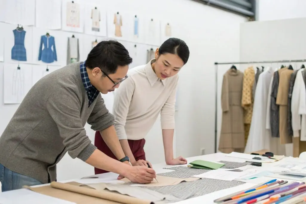 Two Chinese fashion designers intently discussing garment patterns in a studio, surrounded by design sketches